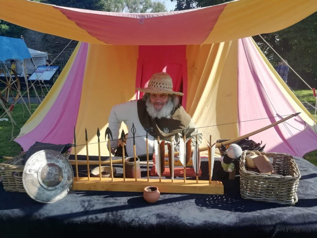 A medieval craftsman sits in front of his tent showing different types of arrowhead